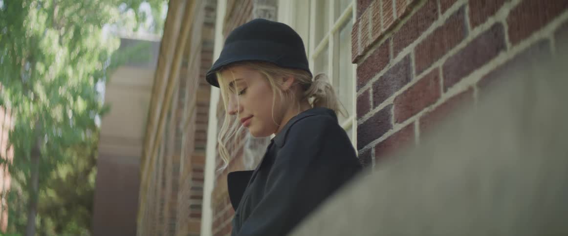 Blonde woman wearing a black hat and coat leans against the brick wall outdoors