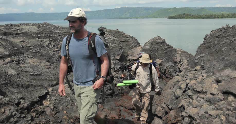 People walk up a rocky hill on a coastline while carrying equipment