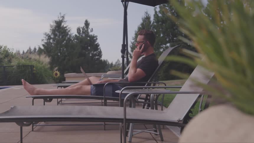 A caucasian male laying on a chair outside talking on a cell phone