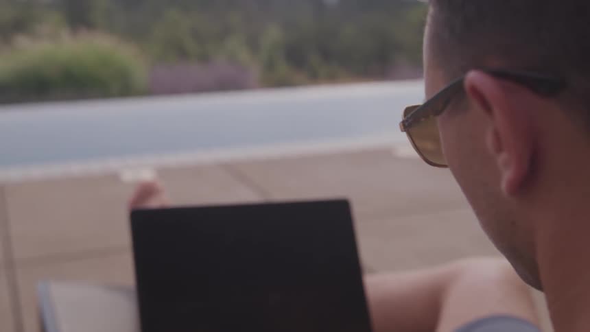 A man sits on a patio chair with legs extended working on a laptop