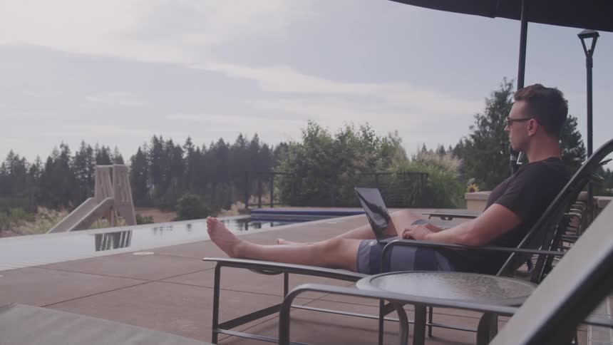 A man sits on a pool deck in a lounger and types on his laptop