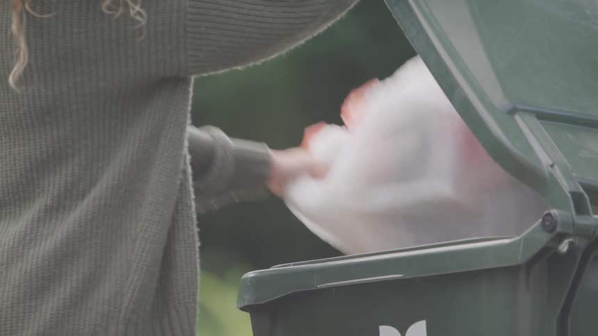 A female in a baggy sweater walks to a garbage can that is outside and places a large plastic garbage bag inside