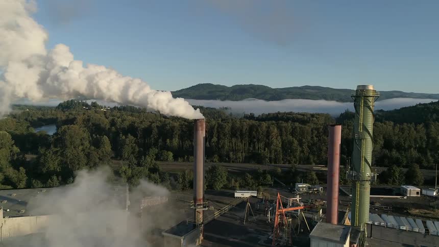 Steam coming out of a stack at a factory surrounded by forests and mountains