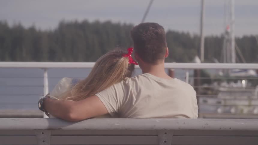 A young couple sits on a bench in front of the water cuddling