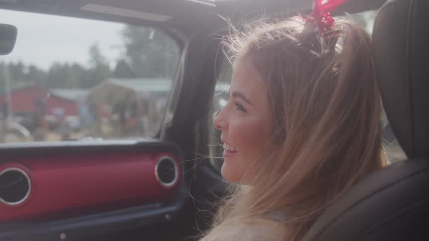 A blonde haired woman turns her head to the driver of the car she is in and laughs