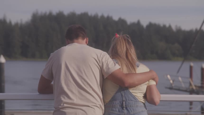 A man has his arm around a woman as they stand at a wall at a marina