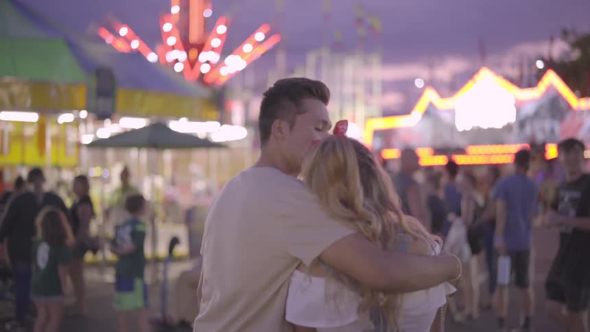 A young male and female embrace while standing within a fairground full of game booths and rides