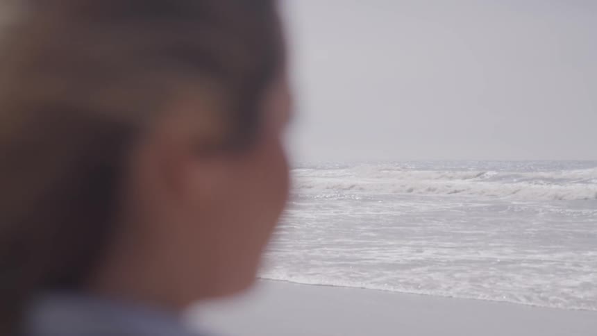 A woman stands at the shore and looks out to sea on a windy day