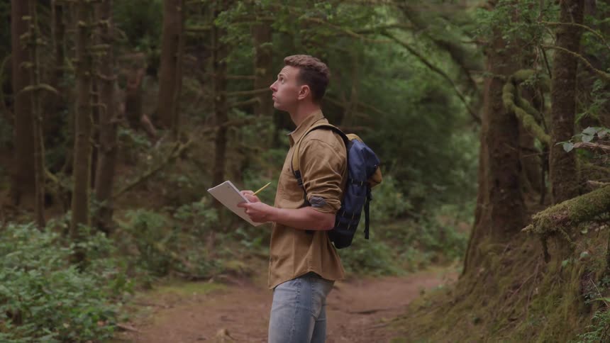 A young man writes in a notebook as he looks up at trees in the forest