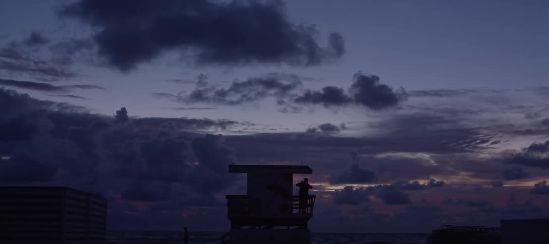 The silhouette of a man in a lifeguard tower against a cloudy sky at sunset