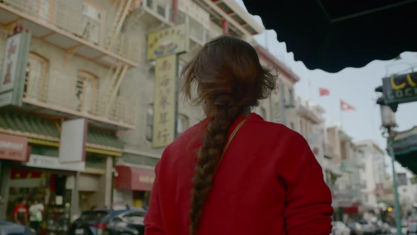 A young woman walks down a city sidewalk and looks up toward the buildings and signs around her