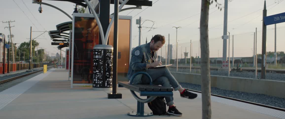 A man sits on a bench at a train depot and uses a tablet