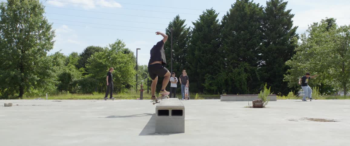 A group of young males skate around an open lot and perform tricks on concrete benches