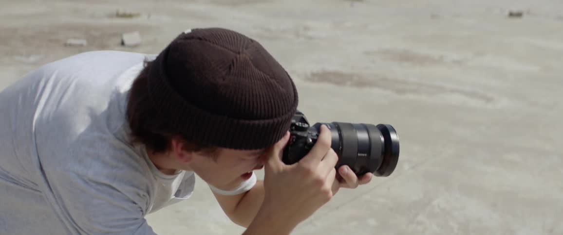 Photographer wearing a brown beanie hat takes picture of people riding a skateboard outdoors