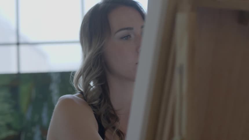 A young woman in an art studio concentrates on painting on a canvas