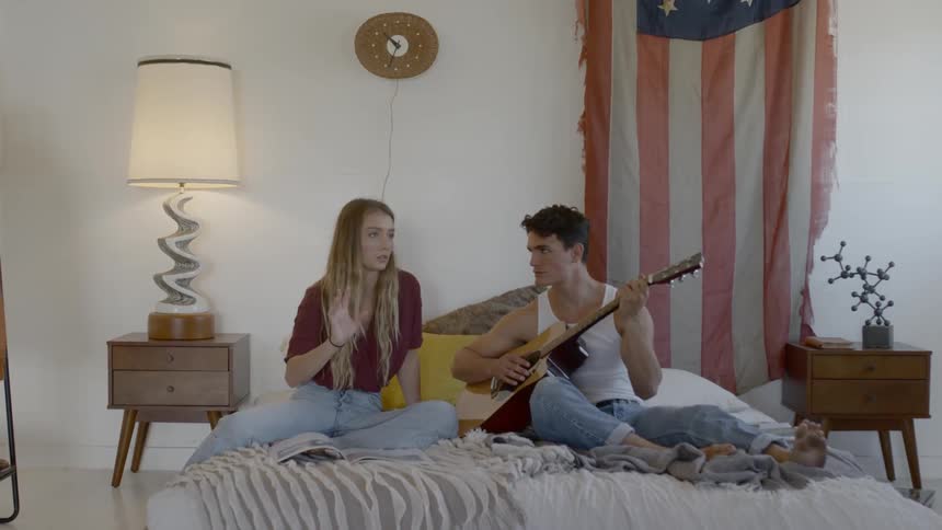 Blonde woman and man playing the guitar sit on the bed before the american flag in the room