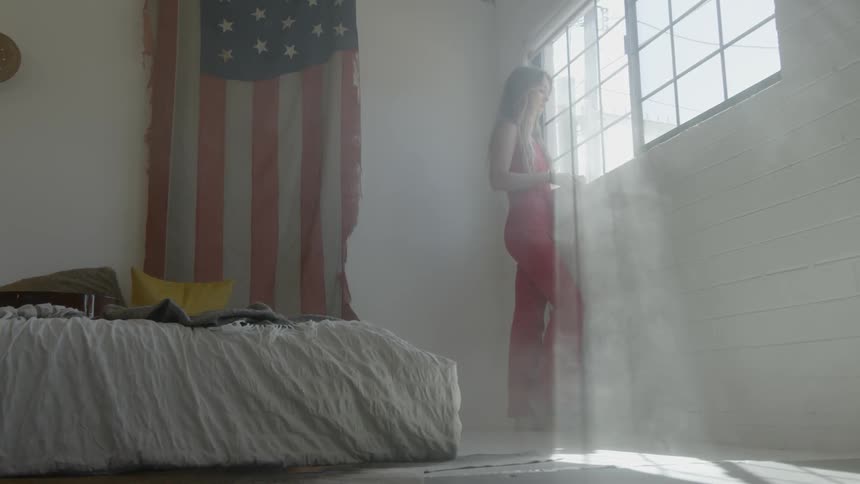 Blonde woman wearing a red dress looks out the window next to the american flag hung on wall in a room