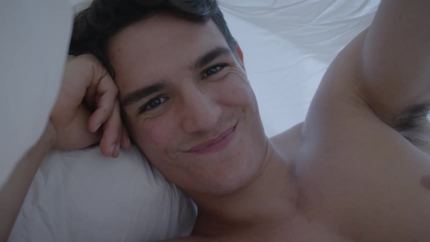 A shirtless man smiles as he holds up the white sheet he is lying under