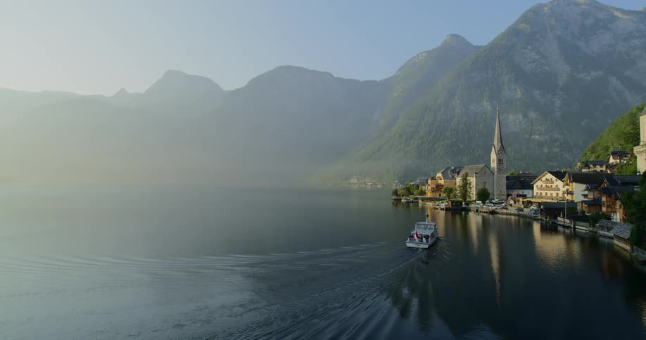A boat travels towards a small town at the edge of a large body of water as a mountain range towers in the background