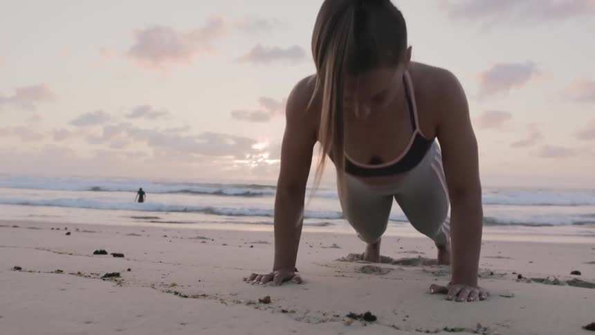 Woman does push ups on beach while surfer moves out from beach
