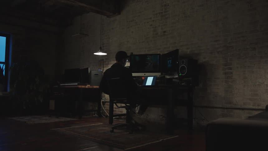 A man sits at his desk with multiple monitors and a tablet in a darkened room