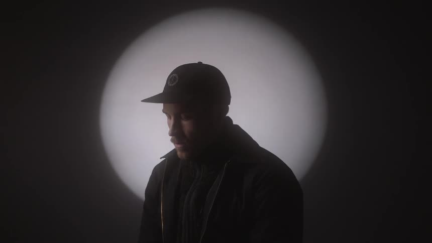 A man sits in a dark bare room with a light shone on him