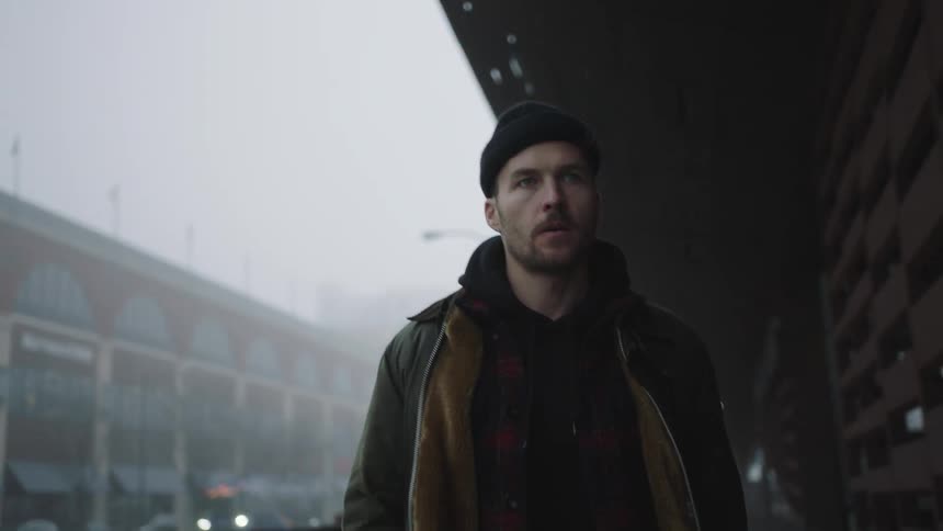A man with a hat and coat walking on a city sidewalk with buildings and vehicles in the background