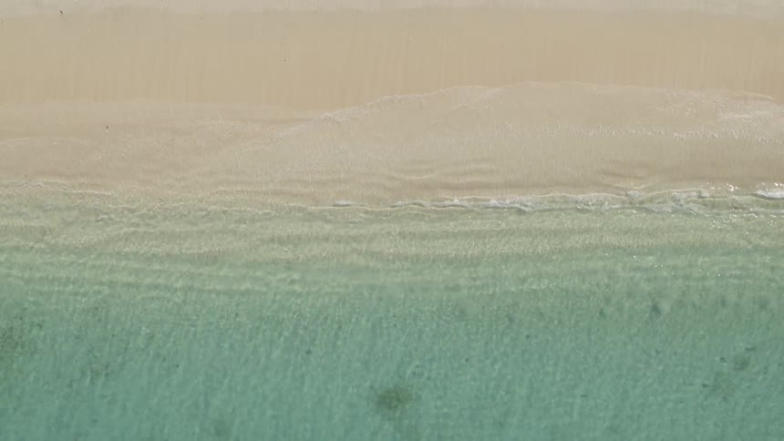 Flying over clear sea water as it forms gentle waves lapping onto a sandy beach