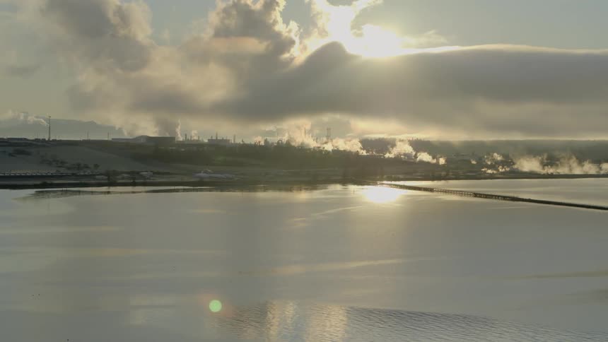 Bright sun backlights an industrial refinery complex along a saltwater bay