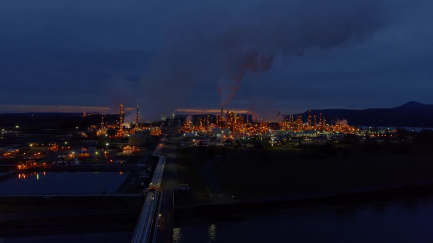 Flying over oil & gas pipes that lead to a large refinery complex at dusk