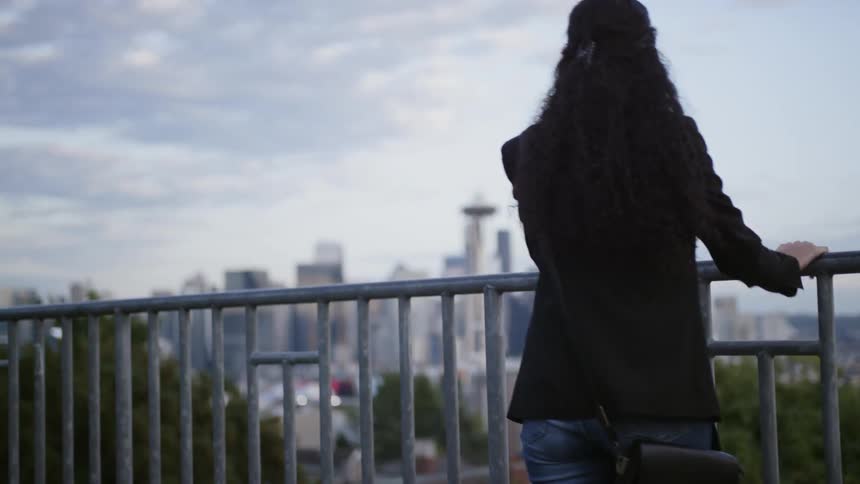 Curly hair woman walks up to the banister and rests her elbows to look at the city view