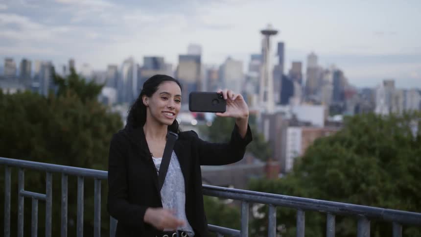 Woman smiles while taking a selfie on smartphone with city downtown as background