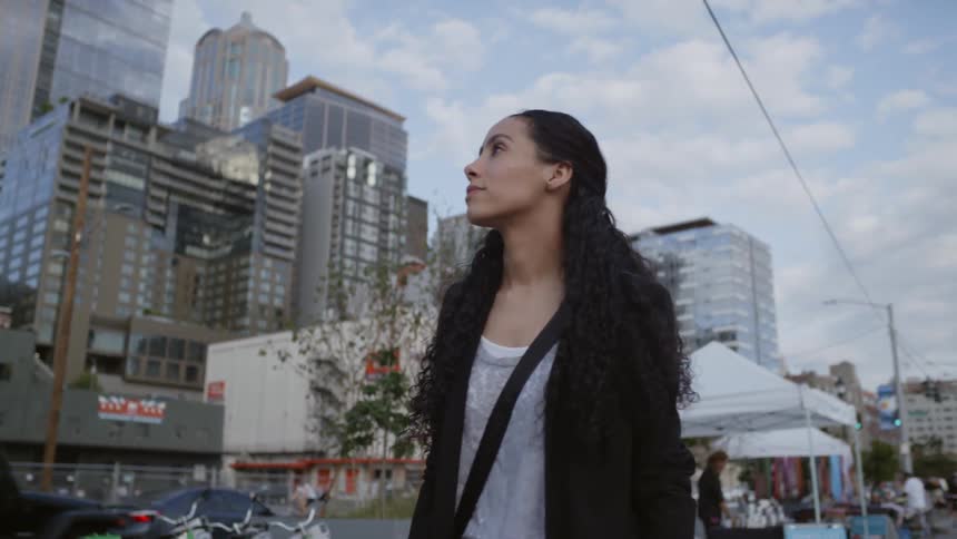 A woman walks a city sidewalk looking towards the buildings