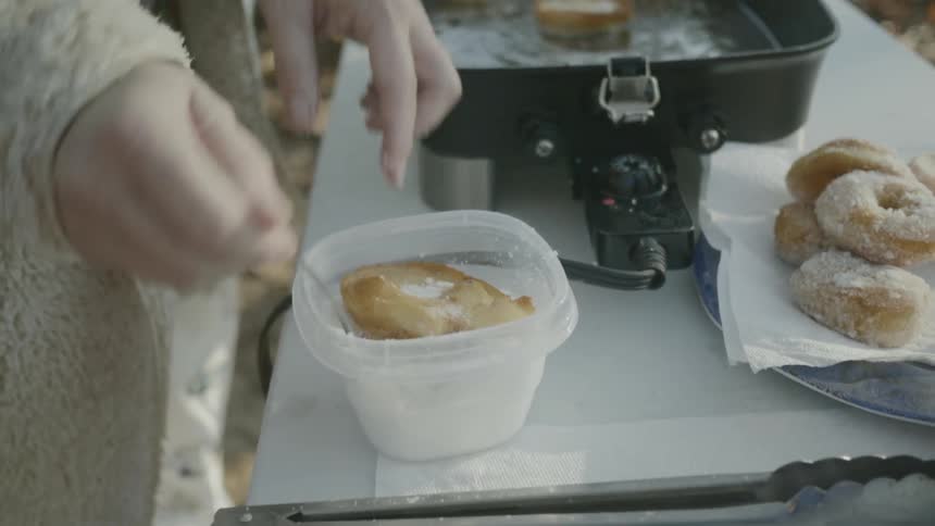 A woman removes a donut from a deep fryer and covers it with powdered sugar