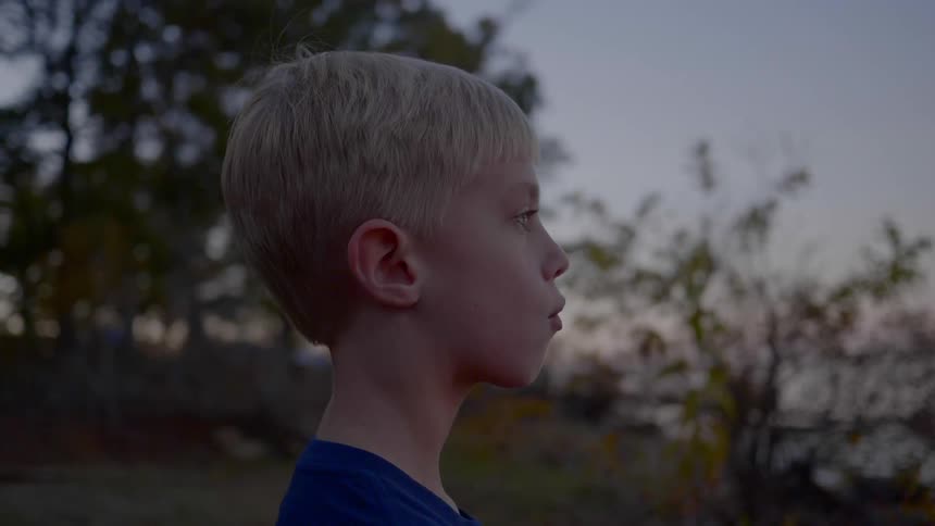Young boy stares ahead as he stands near the trees