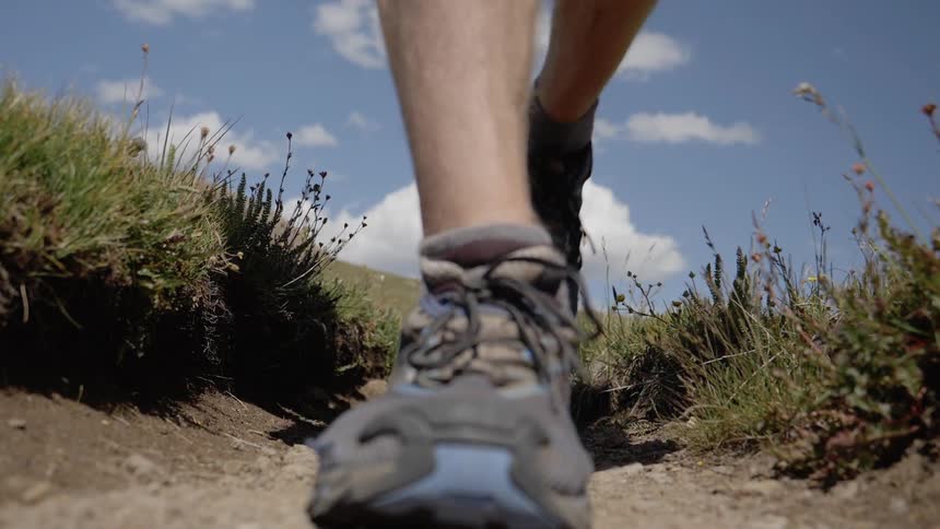 A man in shorts carries hiking poles as he hikes along a dirt trail surrounded by grass