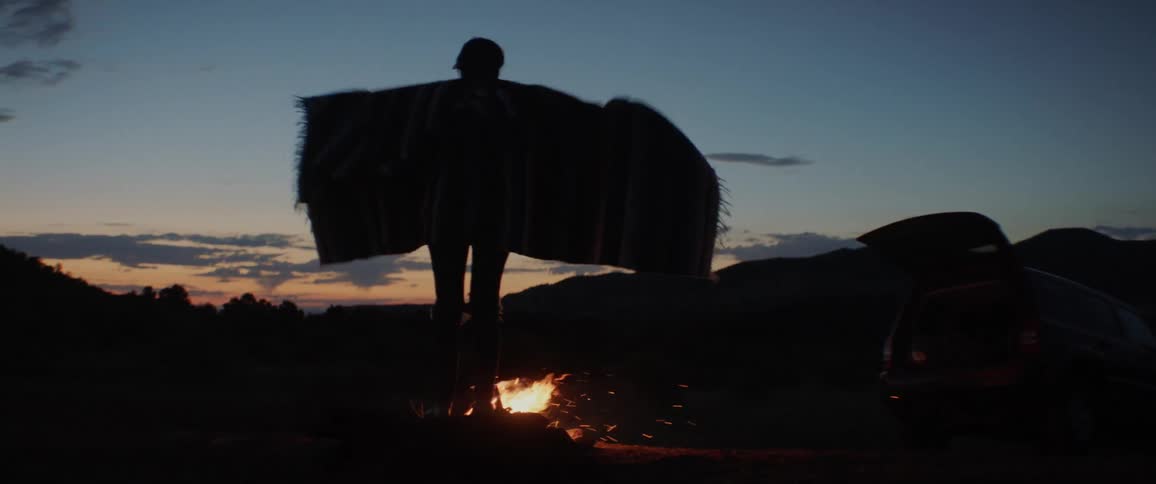 A woman stands waving her blanket around near a fire that burns at night