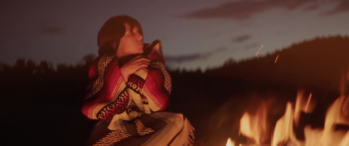 A woman in warm clothing by a fire adjacent to the woods at night