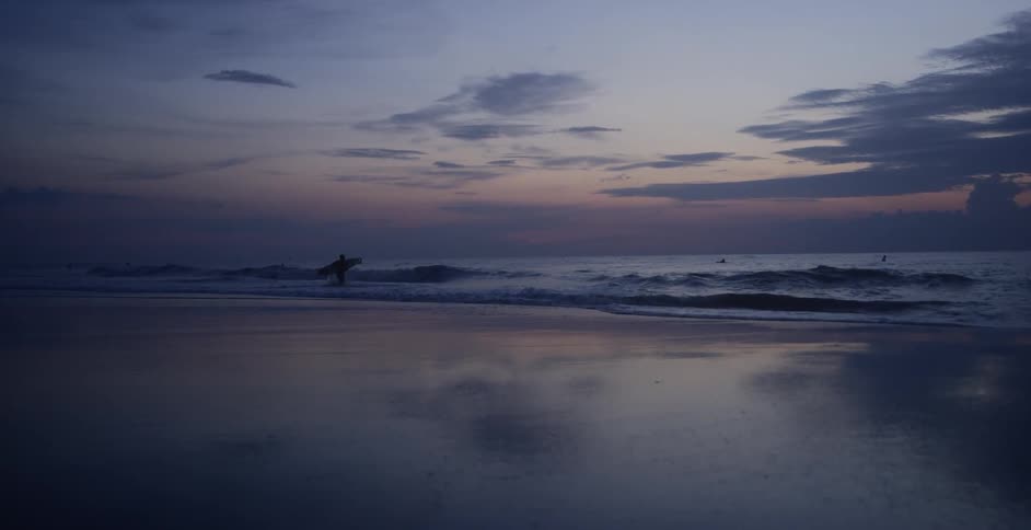 A person carries a surfboard to the ocean coast under a dimly lit sky
