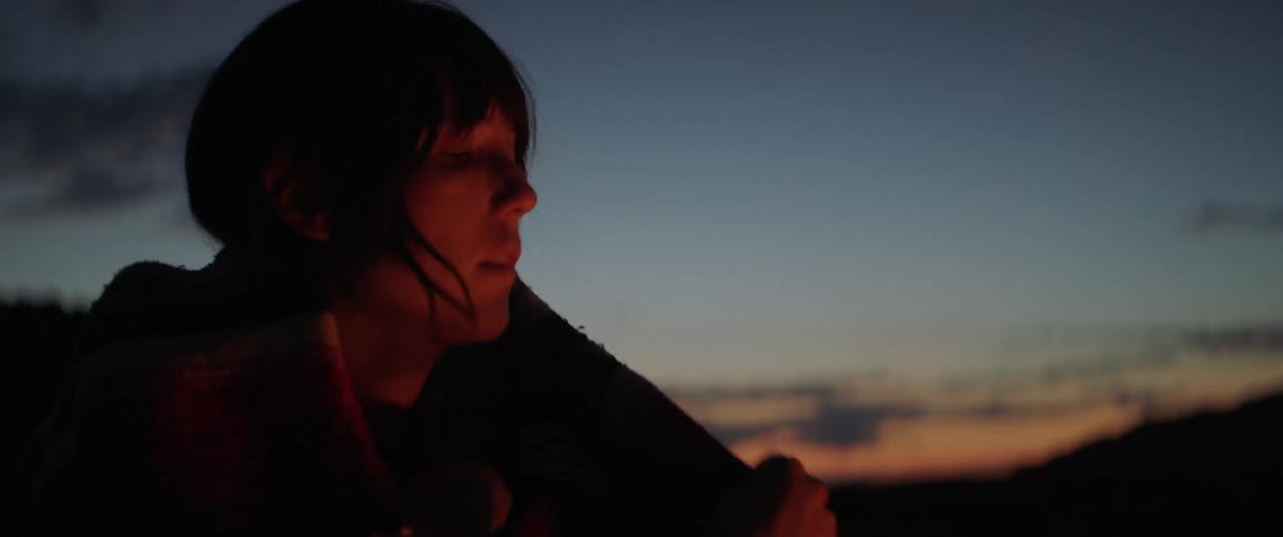 A woman wraps a blanket around her shoulders while sitting by a campfire
