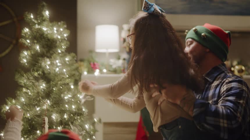 A young girl grabs a christmas ornament and her father lifts her up to put it on the christmas tree