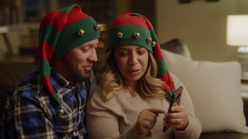 A couple hold up a cell phone to take a selfie together while wearing christmas hats