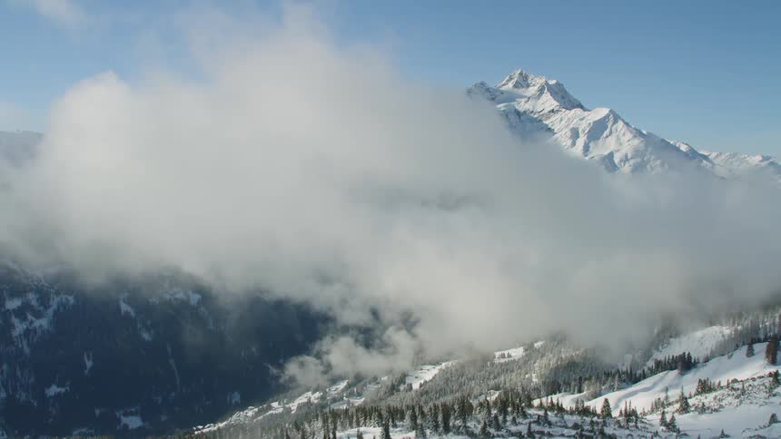 Clouds cover a snow capped mountain covered in evergreen trees