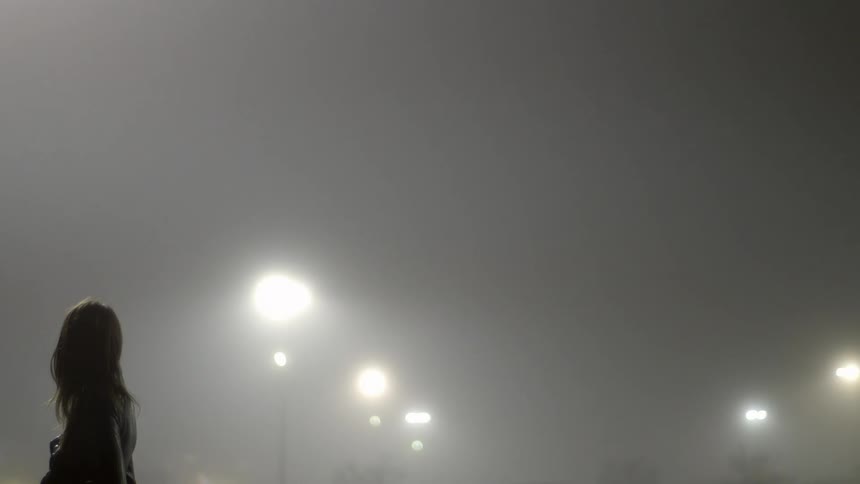 A woman stares to her right while standing alone on a well-lit street on a foggy night