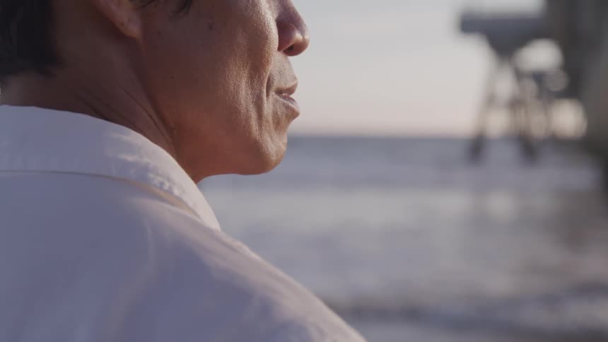 The wind blows a mans shirt collar up as he stands on the beach near the pier