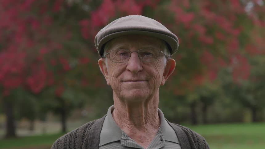 An old white man wearing glasses and a cap smiles while standing outdoors