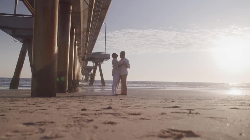 An older couple embracing on a beach under a pier