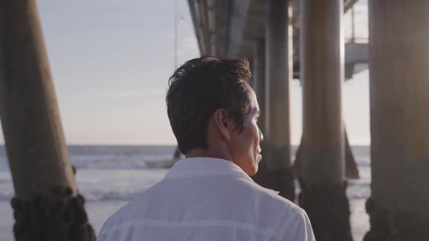 Man standing under pier in beach at sunset next to ocean