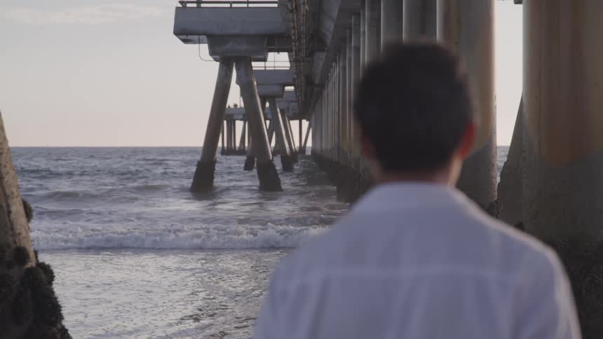 A man stands under a pier and stares at the ocean
