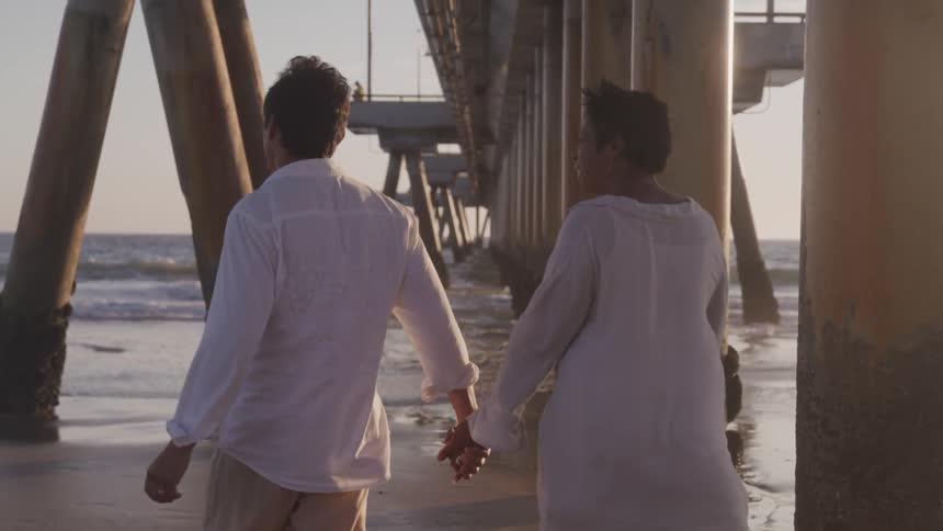 A husband holds his wifes hand as they walk under a pier and laugh